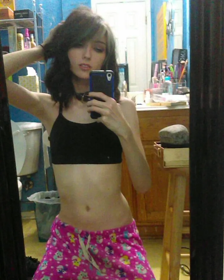 Jeune femme aux cheveux bruns portant un haut noir et un pantalon rose à motifs, prenant un selfie dans un miroir