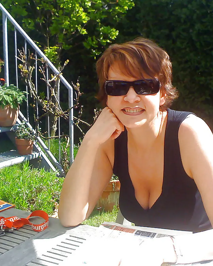 Femme mûre aux cheveux roux portant un haut noir, assise à une table en extérieur sous le soleil
