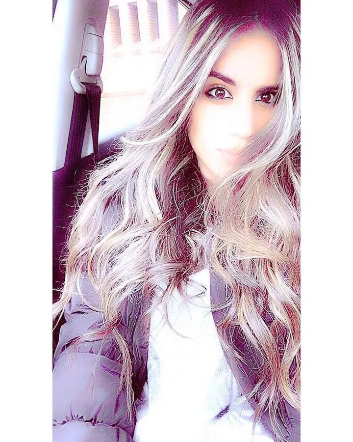 Femme aux cheveux gris, yeux marrons, en photo selfie avec cheveux longs ondulés et veste grise.