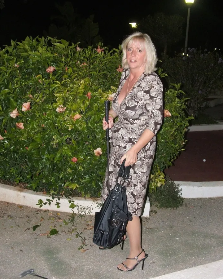 Femme blonde aux cheveux longs, portant un haut noir et un short, posant de profil dans un jardin la nuit.