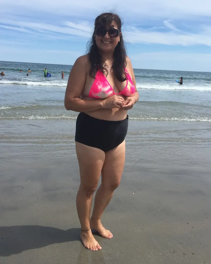 Femme souriante en maillot de bain rose et noir sur la plage, lunettes de soleil, cheveux bruns longs