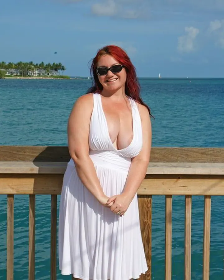 Femme aux cheveux roux, souriante, portant une robe blanche profondément décolletée, debout devant une mer turquoise, sur un ponton en bois.
