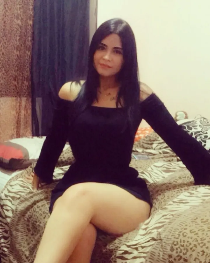 Femme aux cheveux noirs longs, assise sur un lit à motifs léopard, portant une robe noire décolletée, regardant l'appareil photo.