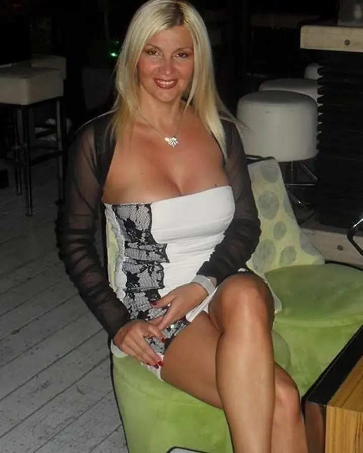 Femme blonde, assise sur un canapé vert, vêtue d'une robe blanche avec un haut noir transparent, souriant à la caméra.