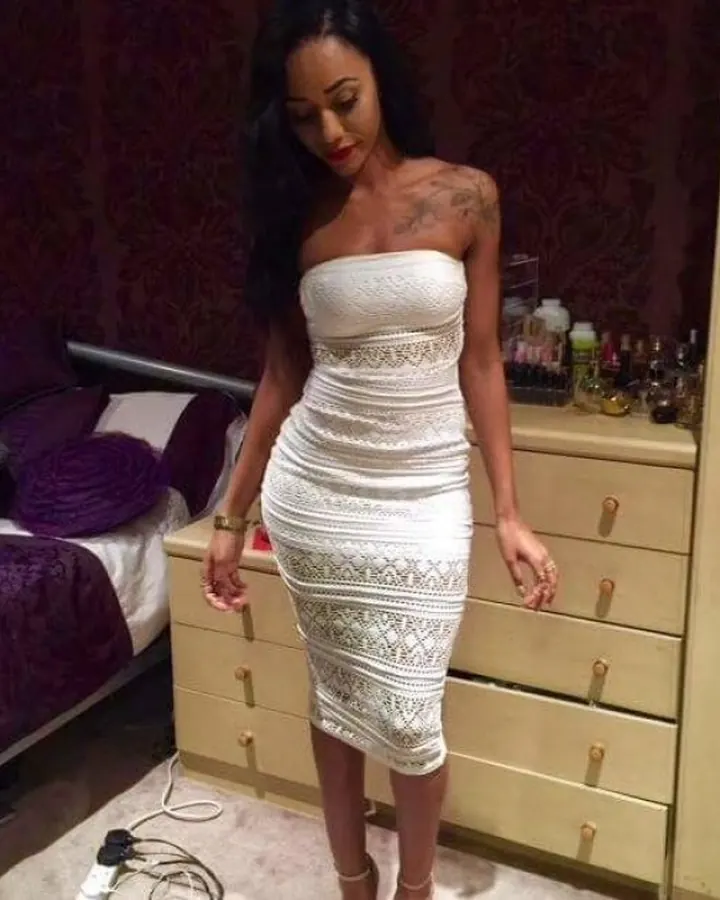 Femme africaine aux cheveux noirs, en robe noire à découpes, posant devant un mur blanc, regardant la caméra.