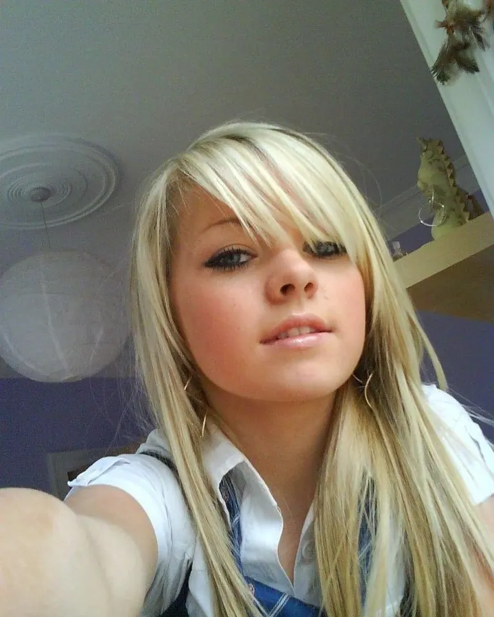 Femme blonde, en haut noir et jeans gris, posant devant un miroir, souriant à la caméra.