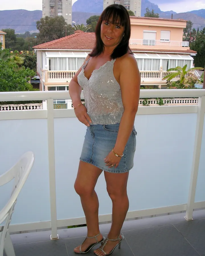 Femme en haut transparent argenté et jupe en jean, debout sur un balcon, mains sur les hanches
