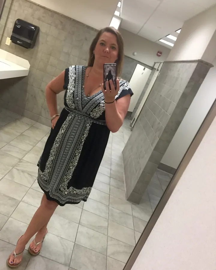 Femme aux cheveux châtains portant une robe noire à motifs, prenant un selfie dans un toilette.
