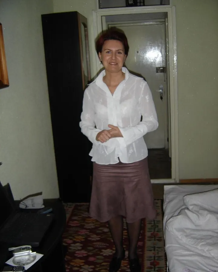 Femme aux cheveux châtains portant une chemise blanche transparente et une jupe marron, debout dans une chambre.