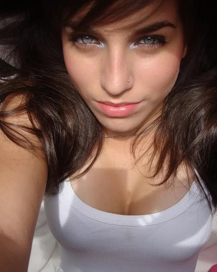 Jeune femme aux cheveux blonds longs, portant un haut blanc à boutons dorés, une jupe blanche courte avec bandes dorées, des collants à mailles roses et des sandales argentées, debout dans une forêt.