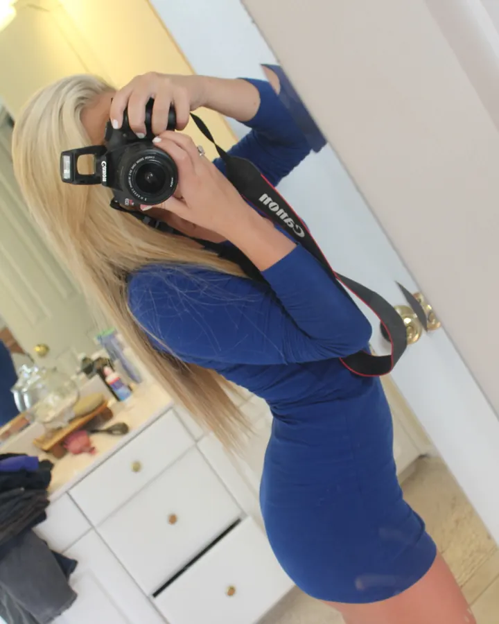 Jeune femme aux cheveux blonds longs, portant une robe bleue ajustée, tenant un appareil photo Canon devant un miroir dans une salle de bain blanche.
