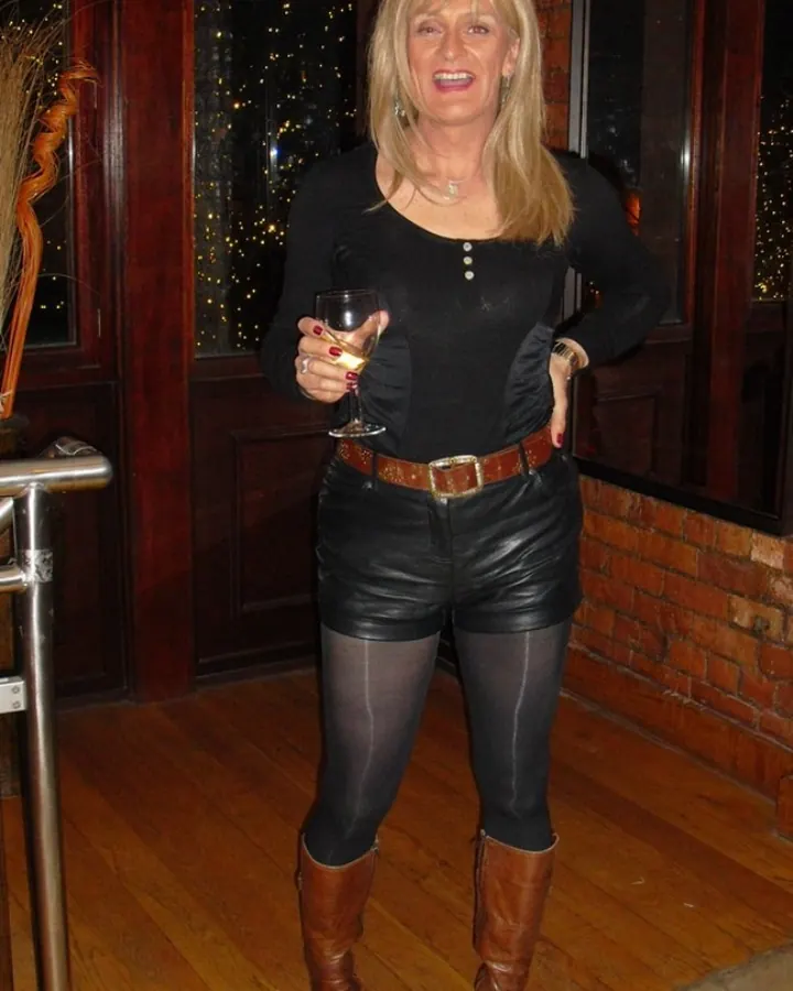 Femme blonde aux cheveux mi-longs, portant un haut noir, une jupe en cuir noir, des collants noirs et des bottes marron, tenant un verre de vin dans une pièce en bois avec des lumières en arrière-plan.