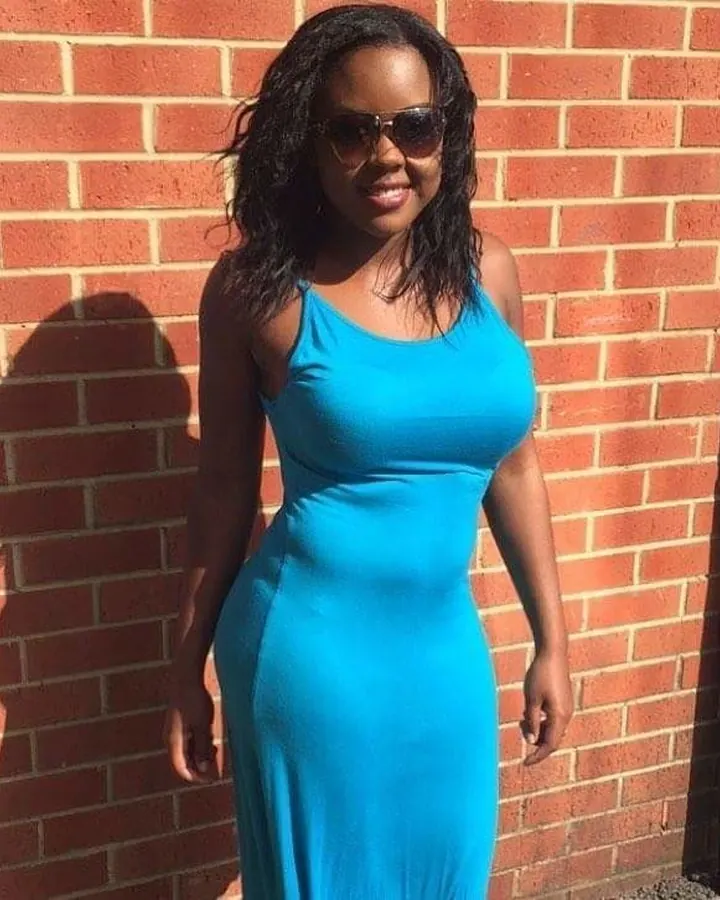 Femme aux cheveux noirs, portant une robe bleue, posant devant un mur de briques.