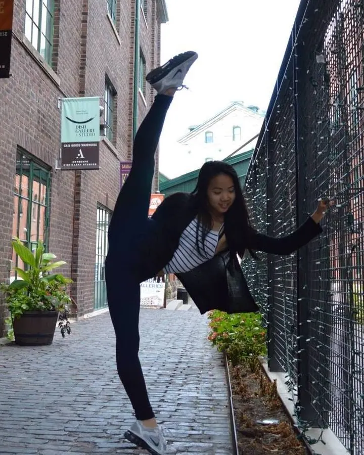 Jeune femme asiatique en haut noir, posant partiellement derrière un mur texturé, avec un regard direct et un haut décolleté.