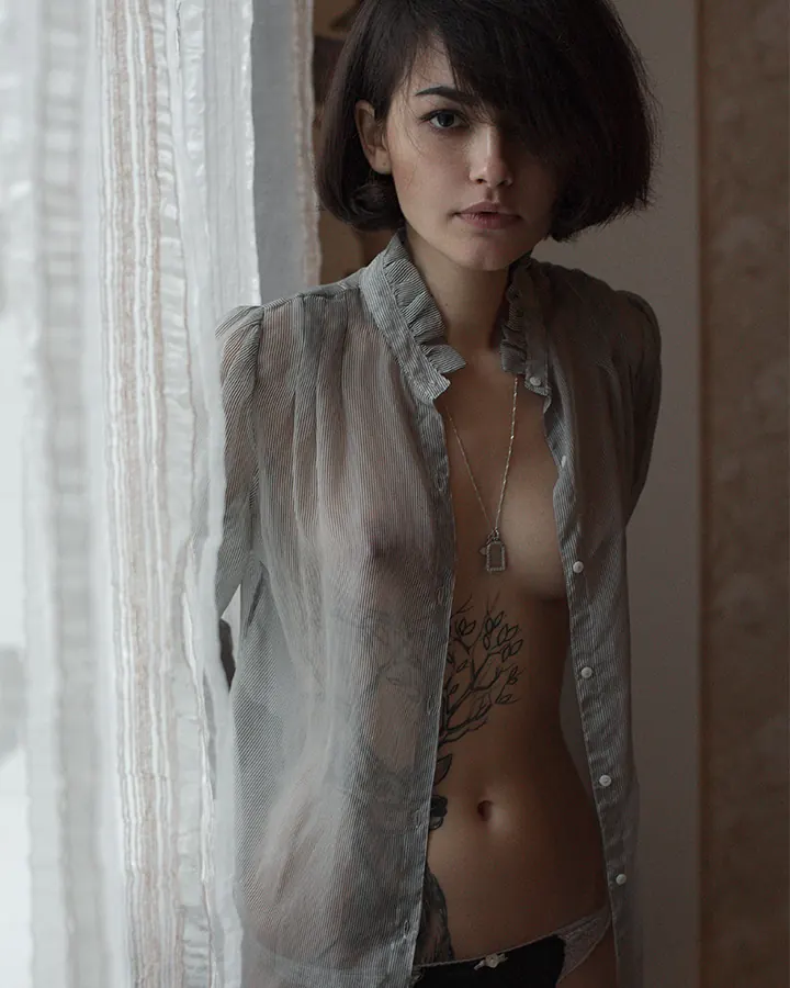 Jeune femme européenne aux cheveux châtains courts, regard intense, vêtue d'une chemise transparente boutonnée, dévoilant un tatouage arborescent sur le torse, devant un rideau blanc.
