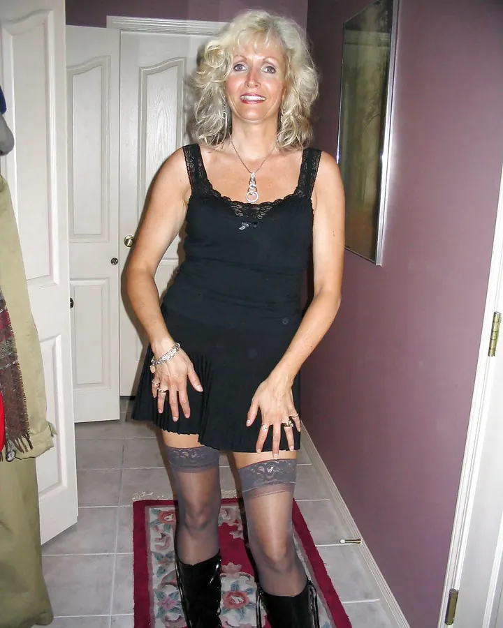 Femme européenne aux cheveux platine, en robe noire, debout dans un couloir, souriant vers l'appareil photo.
