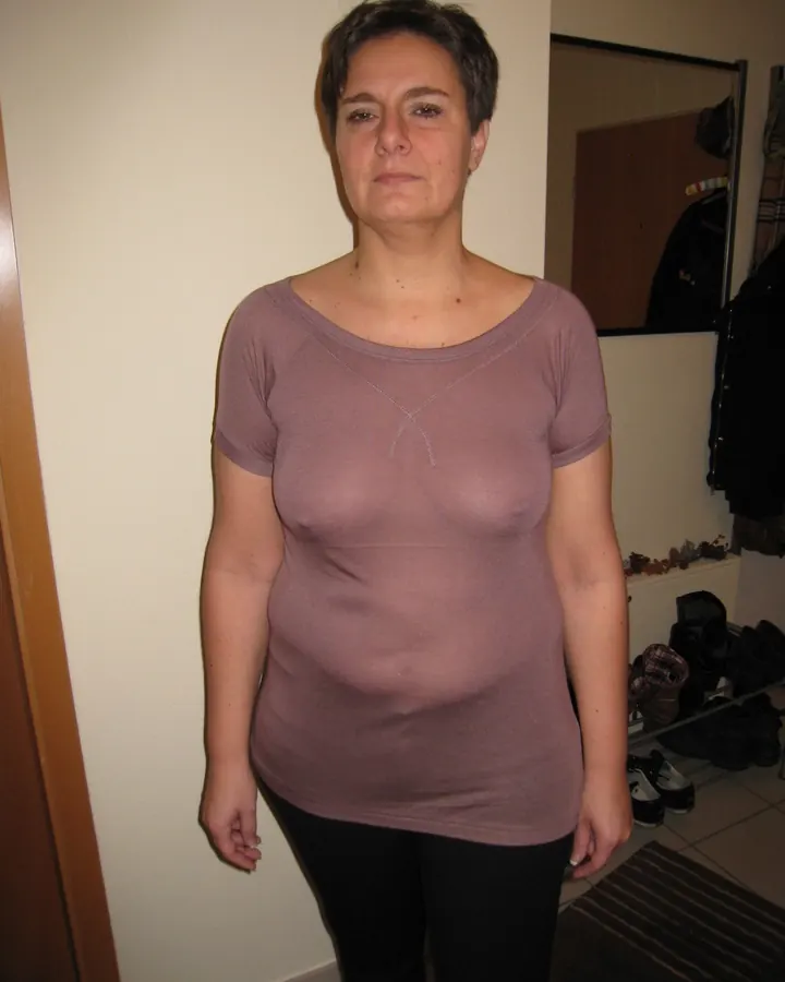 Femme européenne aux cheveux noirs, en robe noire, debout dans un couloir, souriant vers l'appareil photo.