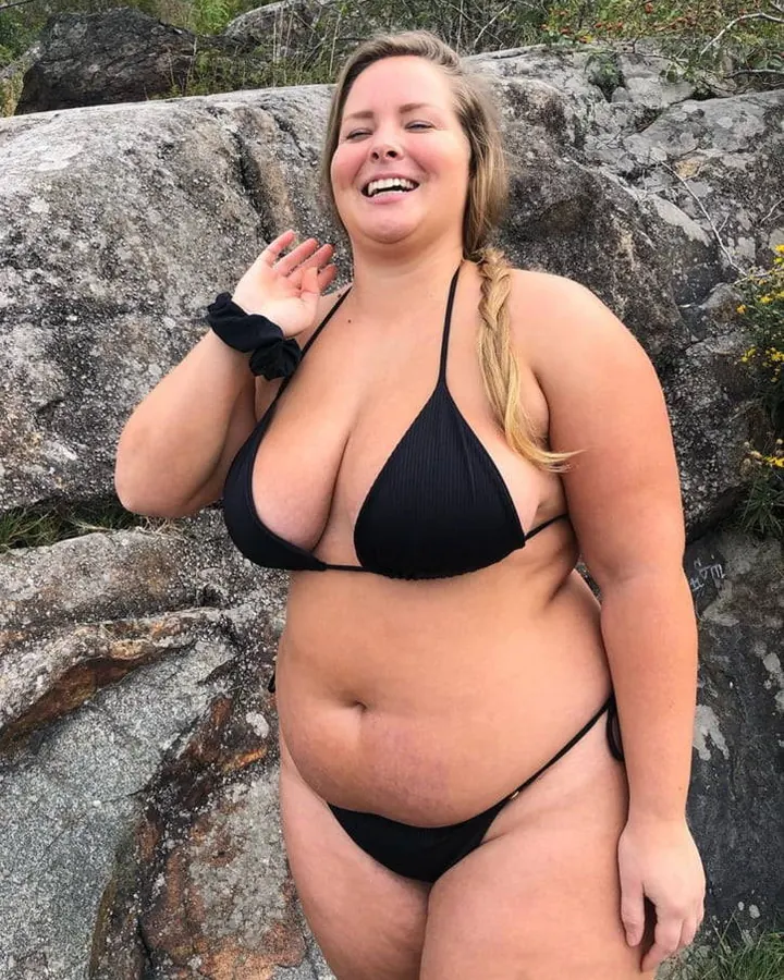 Femme en bikini noir, souriant, main levée, debout près de rochers, en extérieur.