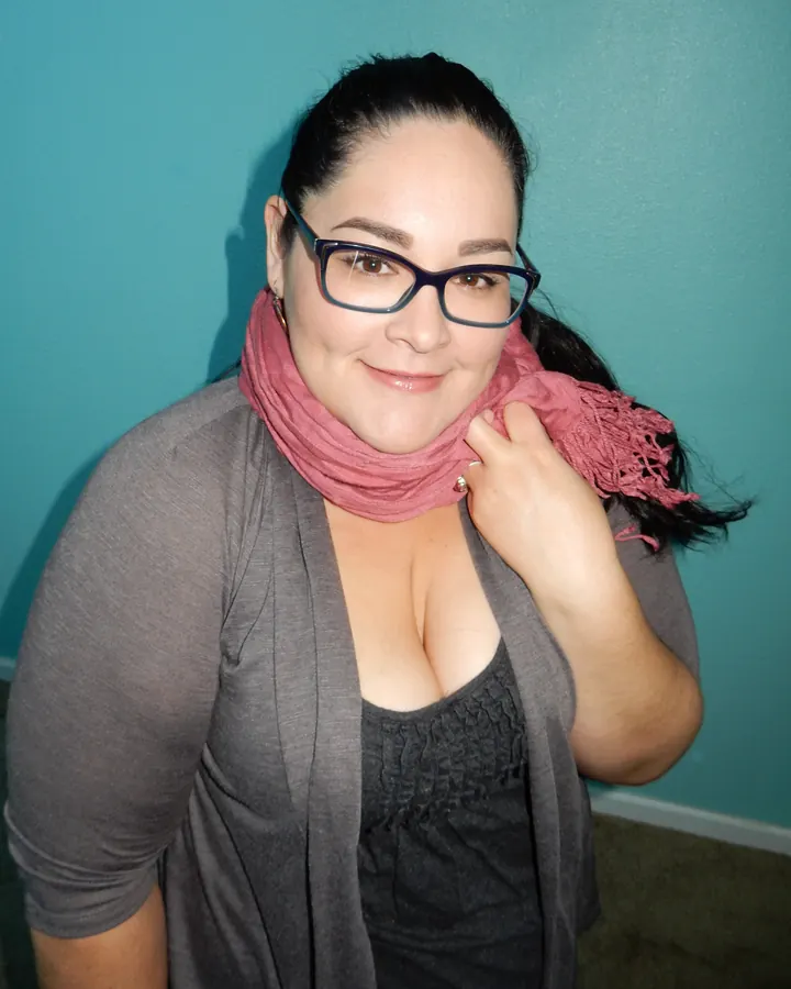 Femme en veste grise et foulard rose, souriant, main sur le foulard, devant un mur bleu.