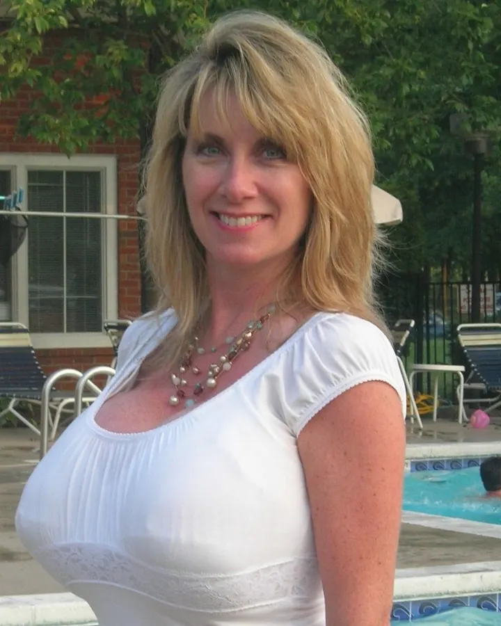Femme blonde en haut blanc, debout près d'une piscine, souriant, regardant l'appareil.