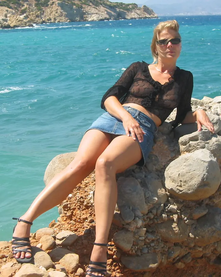 Femme blonde en tenue noire transparente et short en jean, assise sur un mur de pierres au bord de la mer, regardant l'appareil photo.
