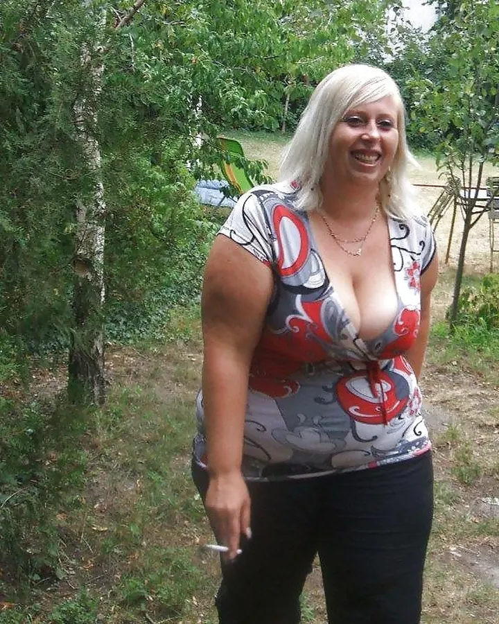 Femme blonde en haut imprimé, pantalon noir, souriante, tenant une cigarette, debout dans un jardin