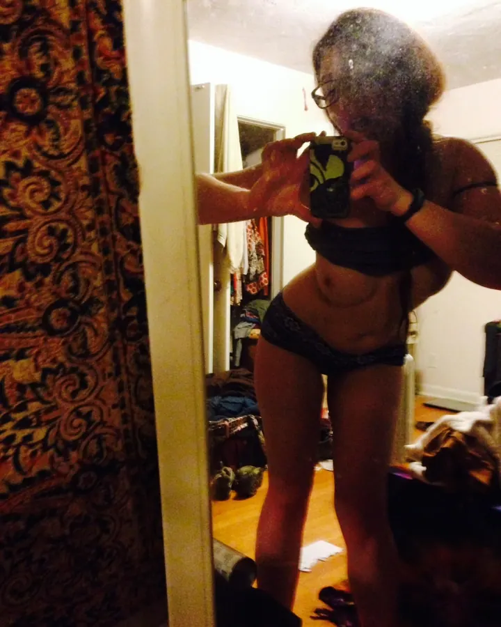 Femme prenant un selfie devant un miroir, portant un soutien-gorge noir et un string assorti, dans une pièce éclairée par une lumière chaude