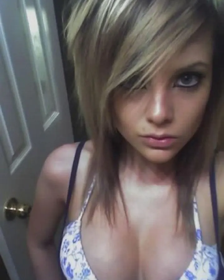 Jeune femme aux cheveux blonds courts, vêtue d'un haut à fines bretelles blanc et bleu, posant près d'une porte blanche.