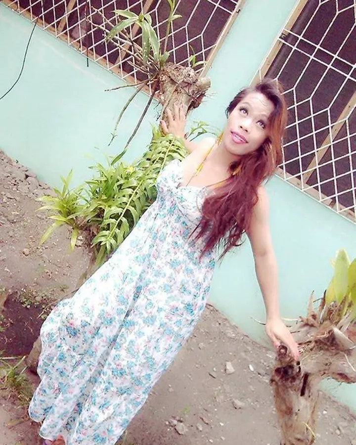 Femme aux cheveux bruns longs, vêtue d'une robe longue à motifs floraux, posant près d'une plante et d'un mur vert clair.
