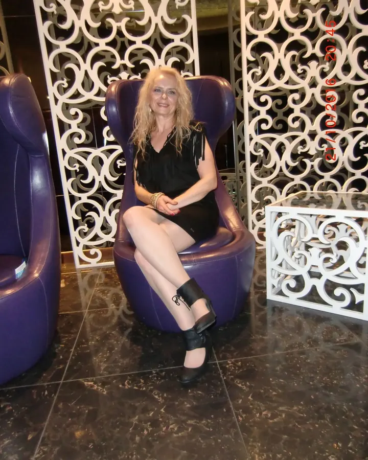 Femme blonde en robe noire assise sur un fauteuil violet dans un intérieur élégant avec des décors blancs.