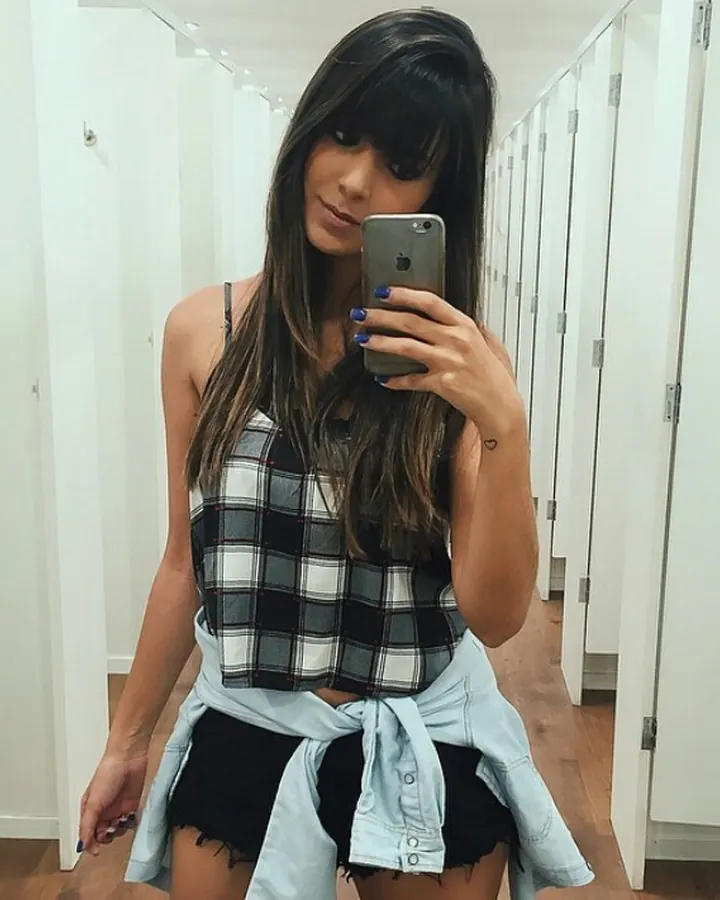 Femme aux cheveux bruns-noirs en haut à carreaux, prenant un selfie dans un couloir blanc