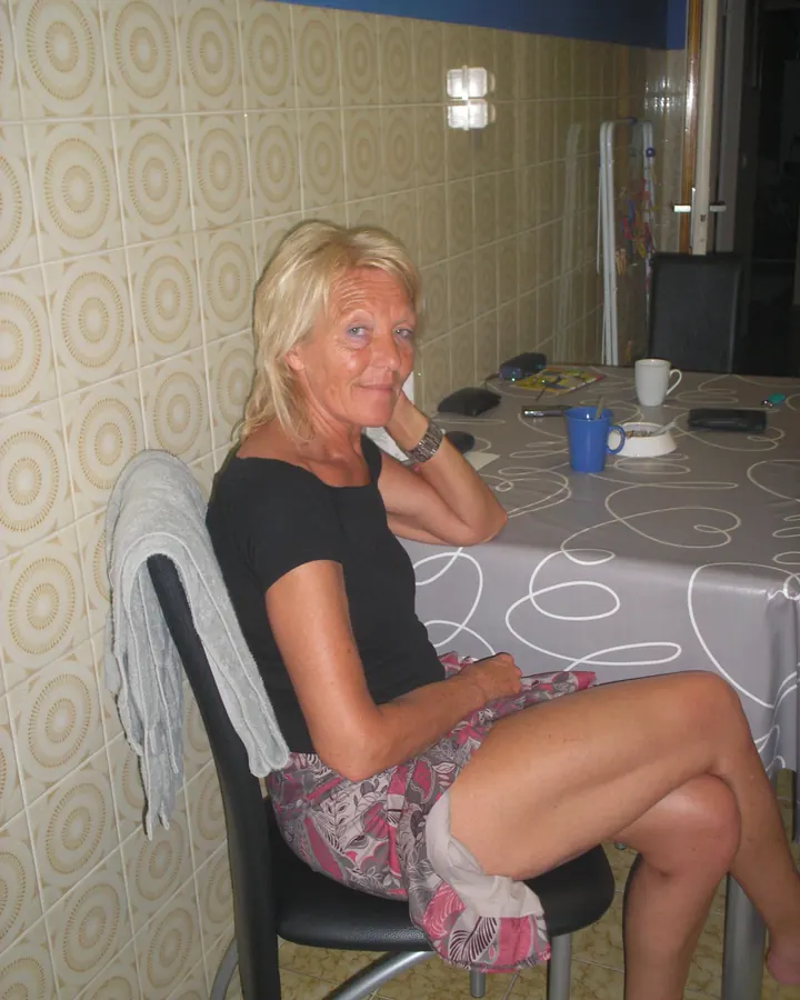 Femme aux cheveux blonds courts, assise à une table, chemise noire, jupe imprimée, sourire chaleureux.