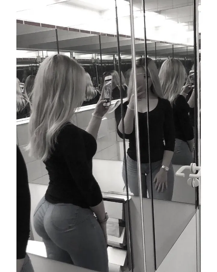 Jeune femme blonde, en tenue noire, prenant un selfie dans un miroir, regard vers l'appareil.