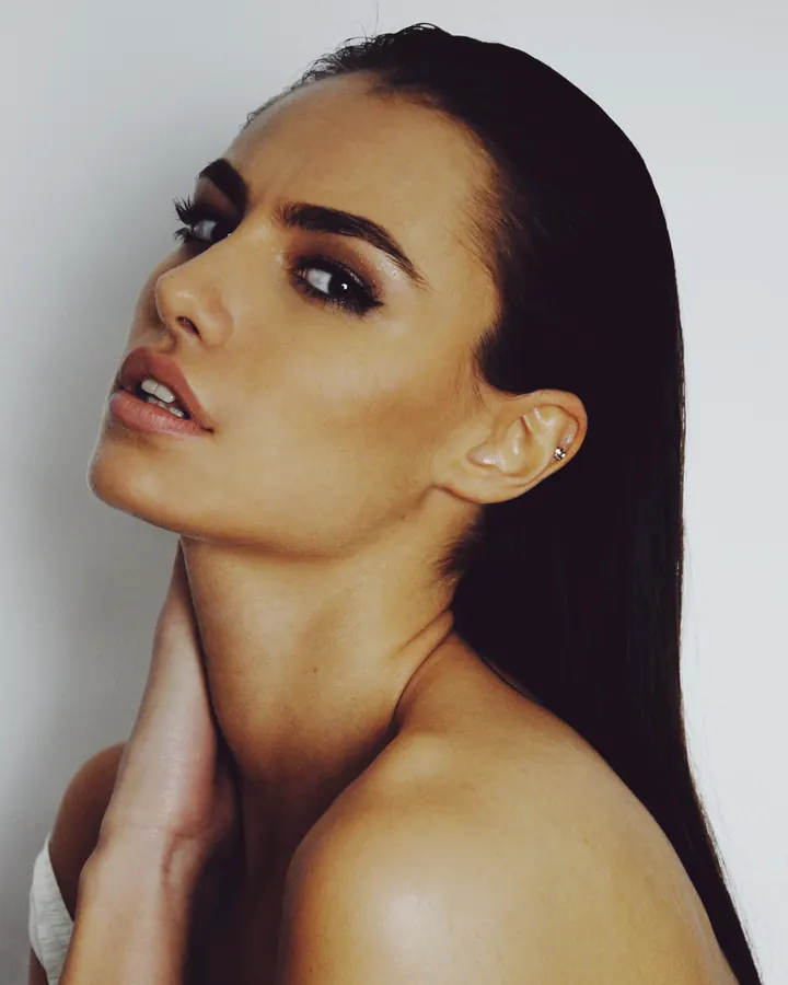 Femme aux cheveux bruns-noirs, regard intense, main sur le cou, peau olive, pose sensuelle contre un mur blanc.