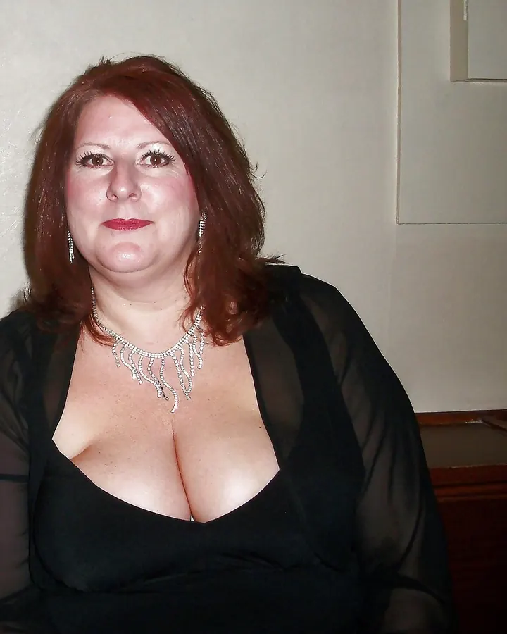 Femme aux cheveux châtains, robe noire transparente, collier, regard direct, peau blanche, poitrine généreuse.