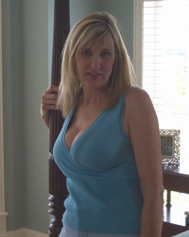 Femme aux cheveux blonds, portant un haut bleu turquoise, debout près d'un escalier en bois, regardant vers l'appareil