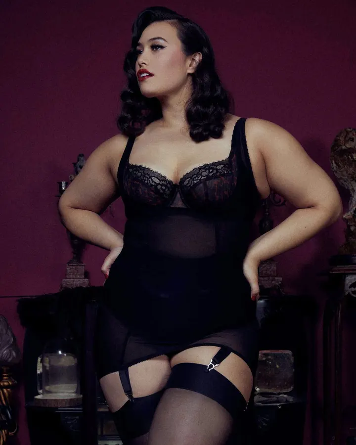 Femme en robe noire, pose sur un lit, mains sur les hanches, regard vers le côté