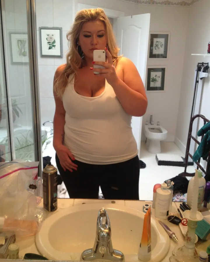 Femme aux cheveux blonds en haut blanc et jupe noire, prenant un selfie dans une salle de bain, regard baissé.
