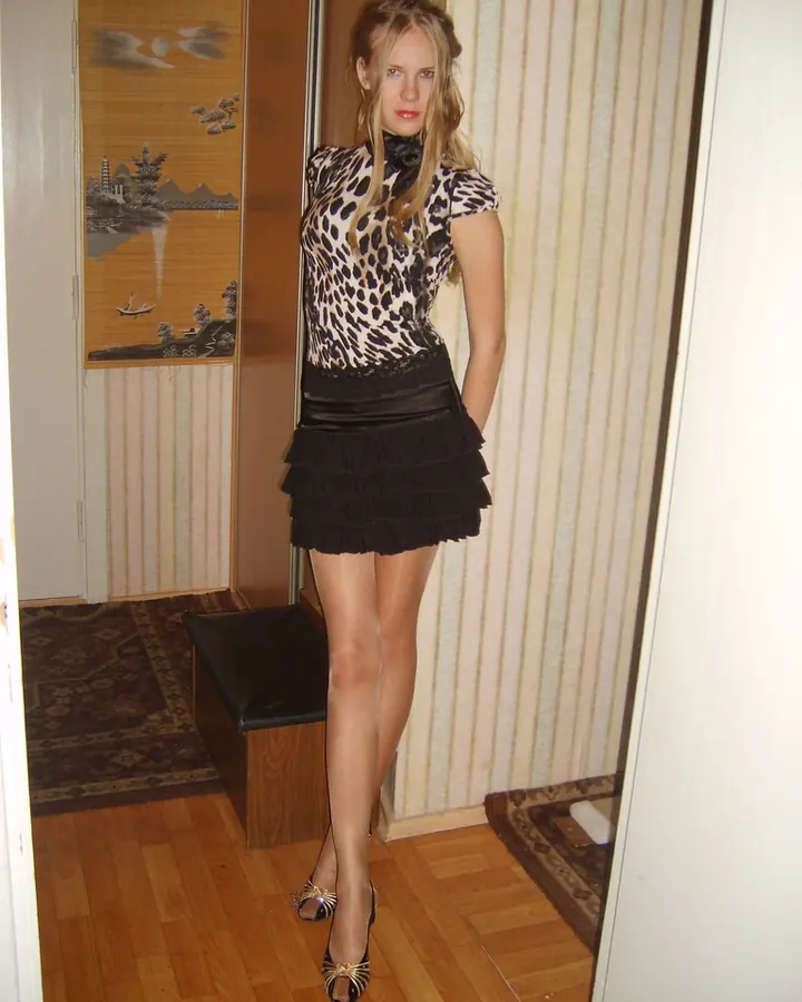 Femme aux cheveux bruns, portant un haut noir, une jupe noire, des collants à mailles et des chaussures à talons, posant dans une chambre.