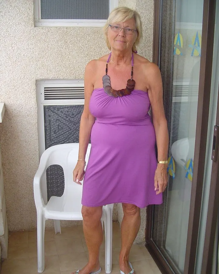 Femme blonde aux cheveux courts, portant une robe violette, debout près d'une chaise blanche sur une terrasse.