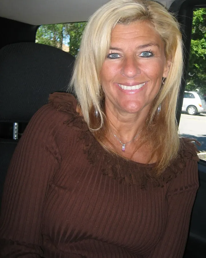 Femme blonde souriante, assise à l'arrière d'une voiture, portant un haut marron à volants
