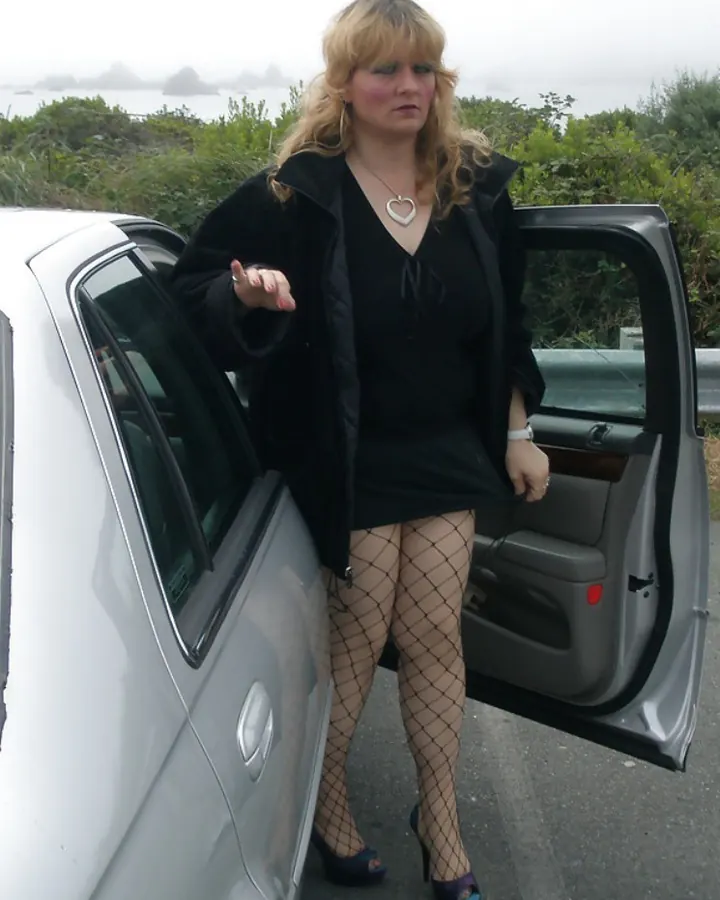 Femme blonde souriante, portant un haut noir à col cœur, debout près d’une voiture blanche, avec des collants à mailles