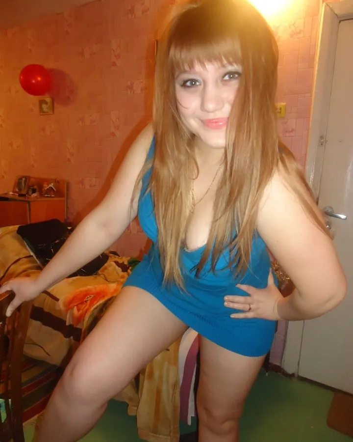 Femme aux cheveux blonds, souriante, portant une robe bleue courte dans une chambre aux murs roses
