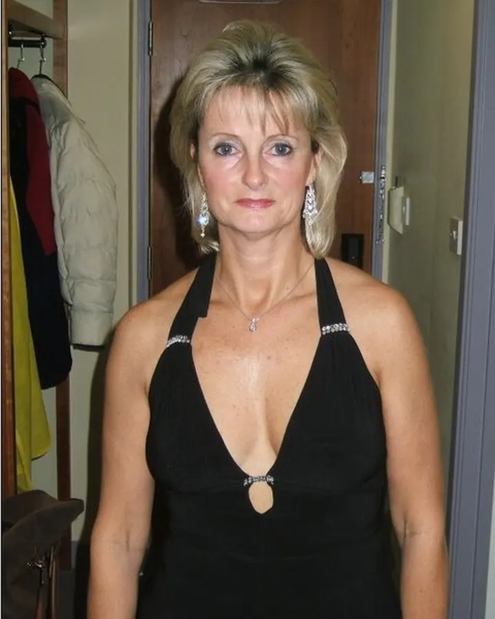 Femme aux cheveux gris platine en robe blanche, assise, tenant un verre de vin, souriante.