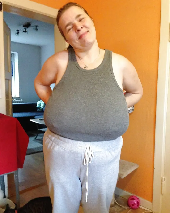 Femme en débardeur gris et pantalon de survêtement gris, souriante dans un intérieur aux murs orange.