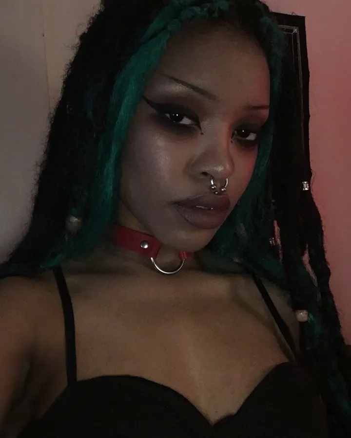 Femme aux cheveux noirs, peau foncée, dreadlocks teintées de vert, collier rouge, regard intense, en tenue sombre.
