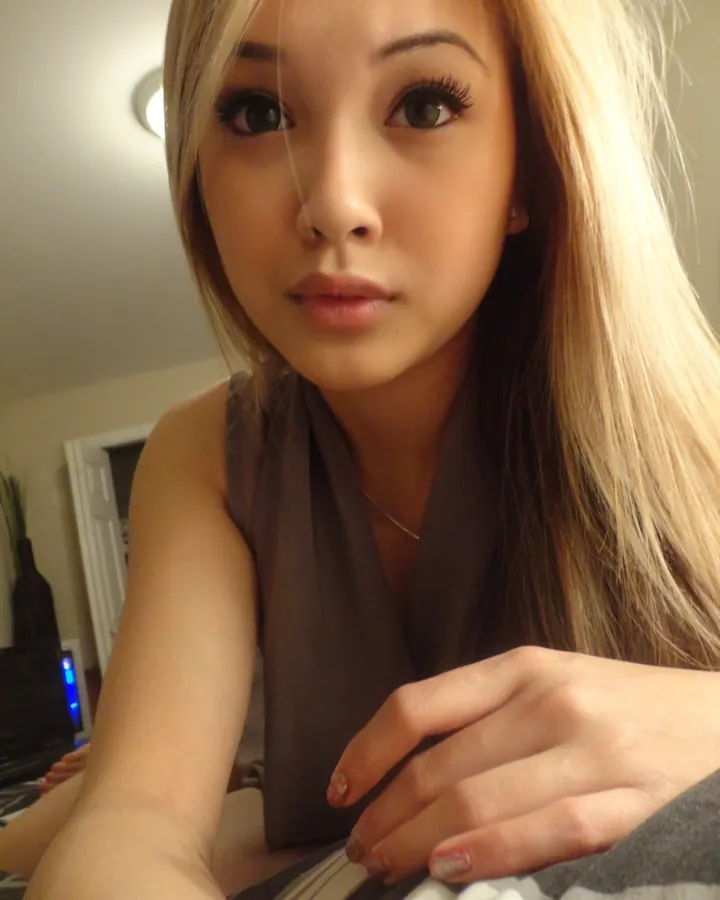 Femme latine, cheveux blonds, yeux marrons, en tenue noire, assise sur une chaise devant une table en verre avec vue sur une terrasse.