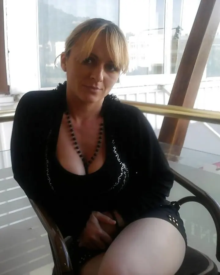 Femme blonde, yeux marrons, en tenue noire, assise sur un canapé devant une fenêtre avec stores verticaux.