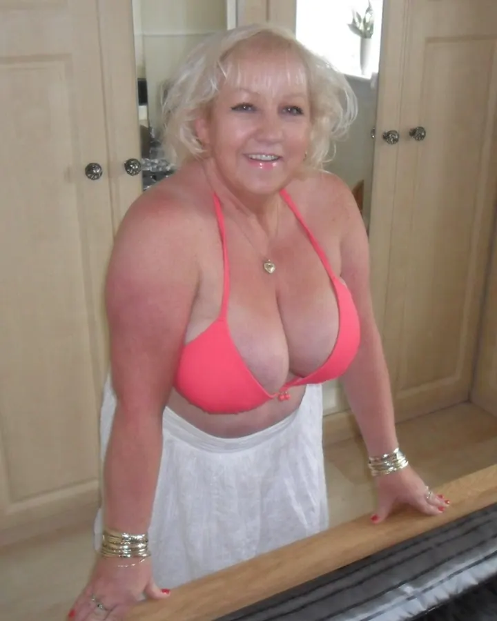 Femme blonde en soutien-gorge rose et jupe blanche, souriante, appuyée sur un lit.