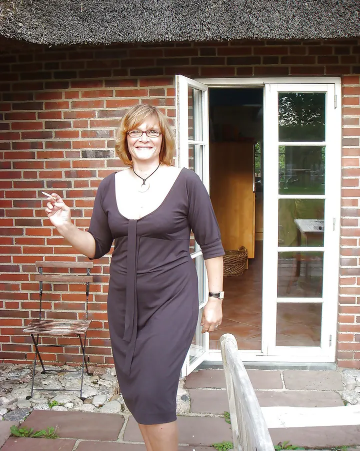 Femme en robe marron, debout devant une maison en briques, tenant une cigarette, souriante, portant des lunettes.